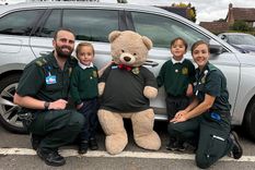 A father and mother with their twins boys and a teddy bear in front of a car