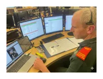An Advanced Paramedic in Critical Care sits at a workstation with four monitors and a laptop, using a video call to support members of the public during a CPR emergency.
