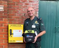 A volunteer in ambulance uniform stands proudly with a defibrillator in front of a case mounted on a wall