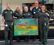 Five members of the Fakenham Community First Responders, three women and two men, stand on a street outside a shop holding an oil painting that has been donated to support fundraising for the group's community work.