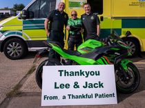 A patient in his motorcycle leathers with two uniformed ambulance staff standing in front of an motorcycle with a cardboard message that says, 