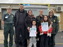 A young boy and girl stand holding framed bravery certificates, with their mother and father behind them, alongside three female ambulance staff in uniform.