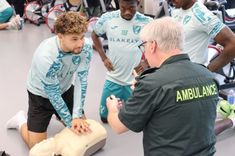 A male footballer player in his training gear is taught CPR on a manikin by a paramedic