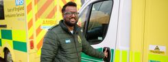 A male paramedic stands beside a marked NHS ambulance, holding the vehicle’s door handle. The ambulance features high-visibility yellow and green markings with East of England Ambulance Service NHS Trust text visible.
