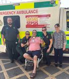 An adult male patient in a wheelchair in the middle of a group with his wife on the far right, a male paramedic on the far left and two female paramedics either side of him