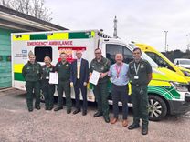 Seven ambulance workers stand in front of ambulance after a commendation presentation, five are wearing green ambulance uniform and one, the CEO of the ambulance service, wears a suit and tie and a manager wears trousers and a shirt