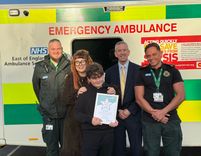 A ten-year-old child stands in front of an emergency ambulance holding a bravery certificate. Behind are a paramedic in uniform, a parent wearing a long coat, a suited Chief Executive, and another uniformed call handlerr.